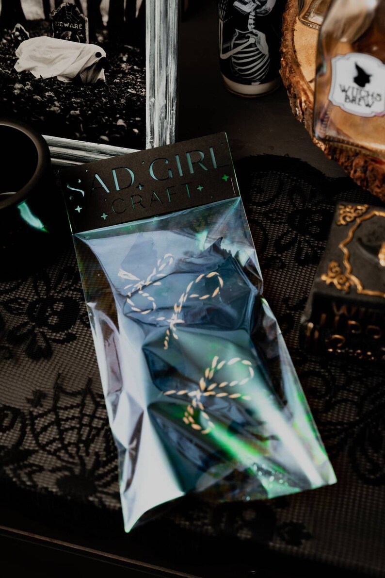 May include: A clear plastic bag with a black card that says "Sad Girl Craft" and contains two gold and black twine bows.