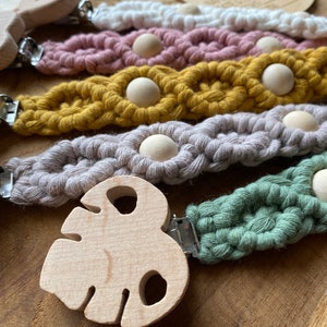 Könnte beinhalten: Fünf gehäkelte Beißringe mit Holzclips und Holzperlen. Die Beißringe sind in verschiedenen Farben, darunter Weiß, Rosa, Gelb, Grau und Grün.