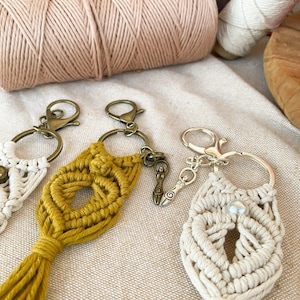 May include: Three handmade macrame keychains with different designs. One keychain is cream colored with a pearl accent, another is mustard yellow with a tassel, and the third is a silver charm with a woman symbol.