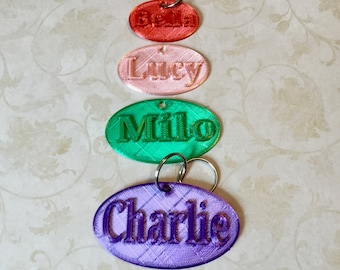May include: Five oval-shaped keychains with different names on them. The keychains are red, pink, green, and purple. The names on the keychains are Bella, Lucy, Milo, and Charlie.