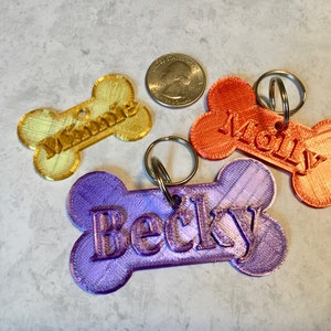 May include: Three bone-shaped dog tags with the names "Winnie", "Molly", and "Becky" engraved on them. The tags are made of different colors: yellow, orange, and purple.