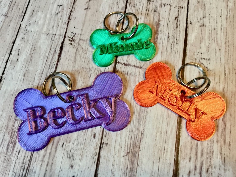 May include: Three colorful bone-shaped dog tags with metal rings. The tags are purple, green, and orange. The names "Becky", "Minnie", and "Molly" are printed on the tags.