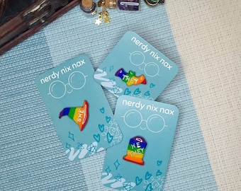 Puede incluir: Tres pines de esmalte coloridos con rayas arcoíris y las palabras "SHE", "THEY" y "HE THEY" sobre un fondo azul. Los pines están en tarjetas con el texto "nerdy nix nax" y un par de gafas.
