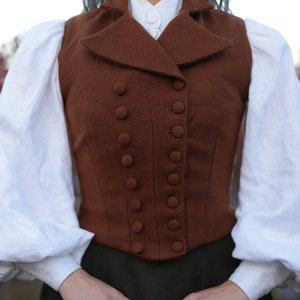 May include: A brown wool waistcoat with a double-breasted button closure. The waistcoat is worn with a white blouse with puffed sleeves and a dark green skirt. The outfit has a vintage or historical aesthetic.