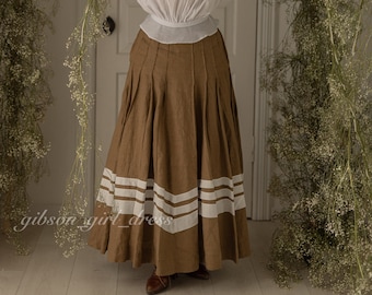 Puede incluir: Una mujer con una blusa blanca y una falda marrón con rayas blancas. Está sosteniendo un sombrero de paja frente a su cara. La falda es una falda larga y la blusa tiene un cuello alto.