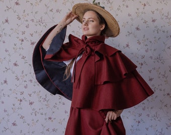 May include: A woman wearing a red dress with a matching red cape and a straw hat. The dress has a ruffled neckline and the cape has a large bow at the neck. The woman is standing in front of a floral wallpaper.