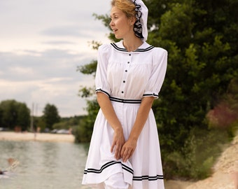 Puede incluir: Una mujer con un vestido blanco con ribetes negros y un sombrero blanco camina por una playa de arena. Lleva medias blancas y zapatos negros.