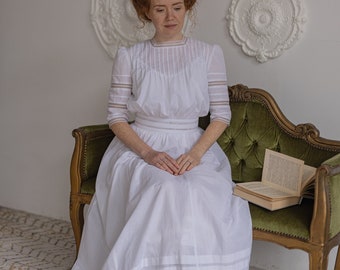 Puede incluir: Una mujer con un vestido blanco con ribete de encaje se sienta en una chaise longue de terciopelo verde. El vestido tiene un escote alto y un corpiño ajustado. La mujer tiene el pelo rojo y mira hacia abajo. La chaise longue es ornamentada y tiene un marco dorado. El fondo es una pared blanca con molduras decorativas.