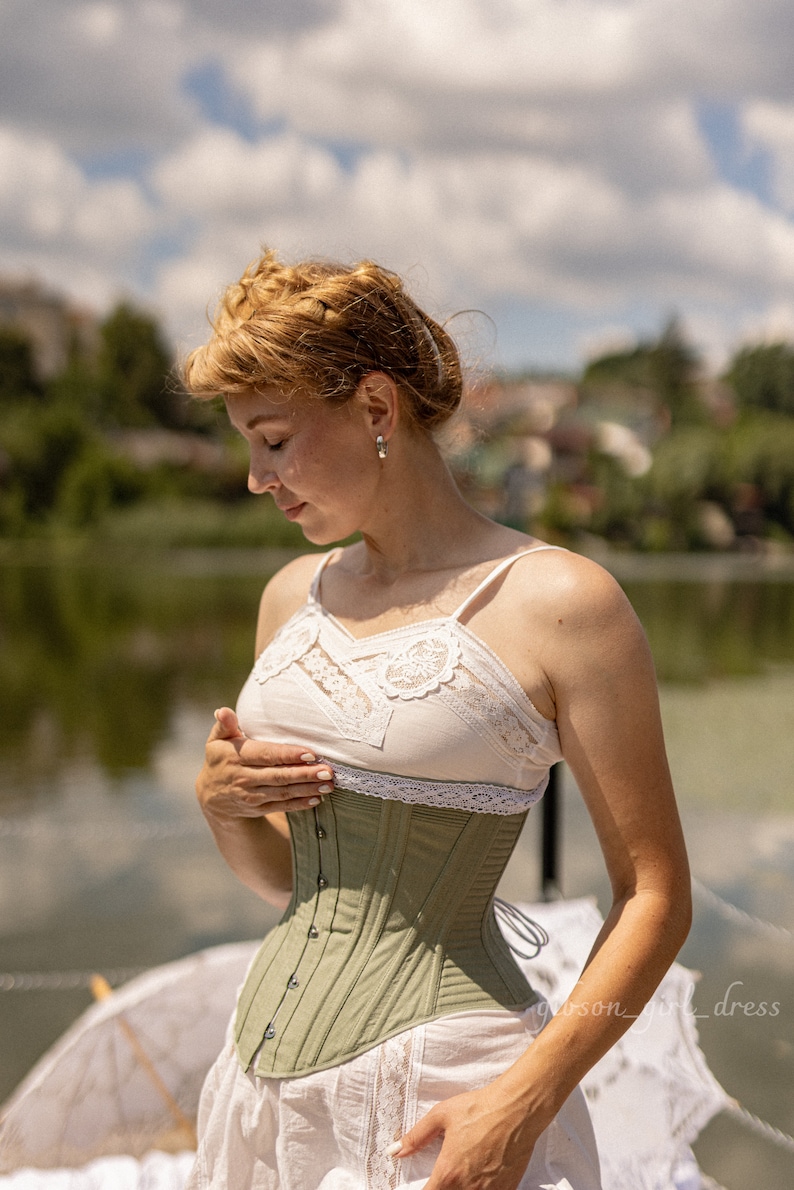 May include: A woman wearing a sage green corset over a white lace top. The corset has a lace trim and is cinched at the waist. She is standing in front of a body of water with a cloudy sky in the background.