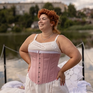 May include: A woman wearing a pink corset with a white lace trim and a white skirt with lace trim. She is standing on a wooden dock with a body of water behind her.