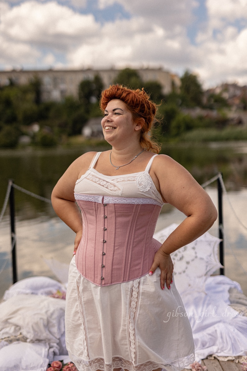 May include: A woman wearing a pink corset with a white lace trim and a white skirt with lace trim. She is standing on a wooden dock with a body of water behind her.