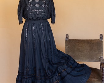 May include: A black vintage dress with intricate lace detailing. The dress features a high neckline, short sleeves, and a long, flowing skirt. The bodice and skirt are adorned with delicate lace accents, creating a classic and elegant look. The dress is displayed on a mannequin.