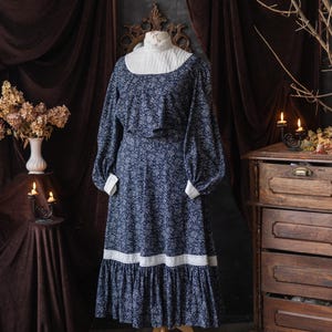 May include: A navy blue floral print dress with long sleeves and a white collar and cuffs. The dress has a ruffled hem and is displayed on a wooden dress form. The dress is a vintage style.