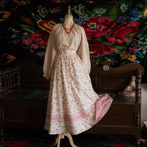 Peut inclure: Une blouse blanche à manches longues et une jupe à imprimé floral sur un mannequin en bois. La jupe est ornée d'une bordure rose et est exposée sur un tapis à motifs colorés.