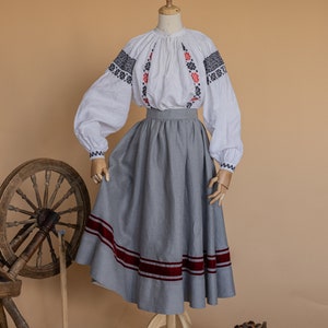 May include: A white embroidered blouse with a grey skirt with red trim. The skirt is on a wooden stand. A spinning wheel and a basket of bread are in the background.