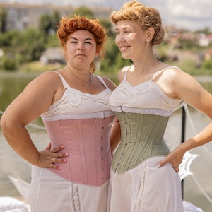 May include: Two women wearing white lace-trimmed slips and corsets. The woman on the left is wearing a pink corset, and the woman on the right is wearing a green corset.