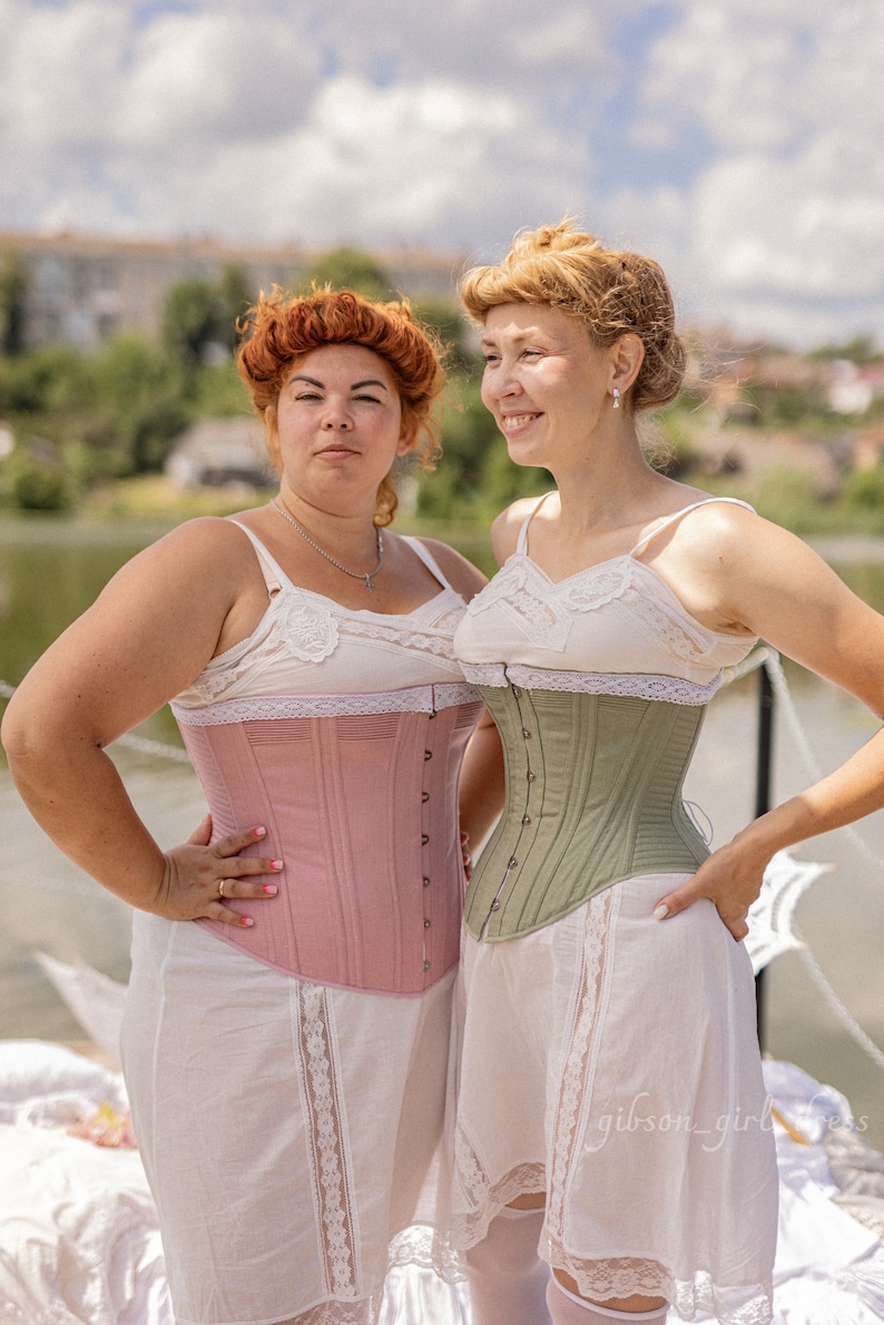 May include: Two women wearing white lace-trimmed slips and corsets. The woman on the left is wearing a pink corset, and the woman on the right is wearing a green corset.