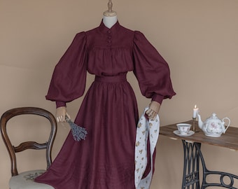 May include: A maroon linen dress with a high neck and long sleeves. The dress has a fitted bodice and a full skirt. The dress is displayed on a mannequin with a white floral sash.