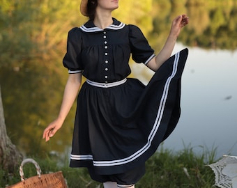 May include: A woman wearing a straw hat and a navy blue dress with white trim stands on a plaid picnic blanket. She is holding a wicker basket and has a white parasol in the background.