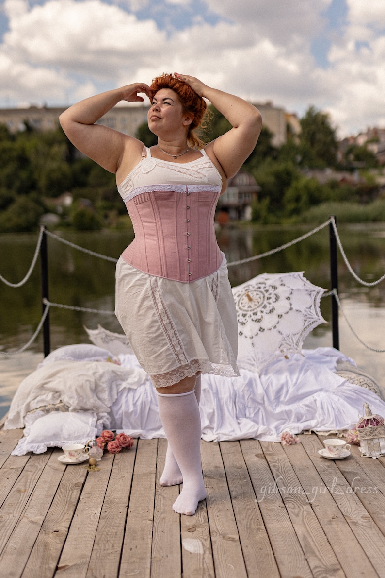 May include: A woman wearing a pink corset and a white skirt with lace trim stands on a wooden dock. She is wearing white knee-high socks and has her hands on her head. There is a white lace parasol and a white blanket behind her.