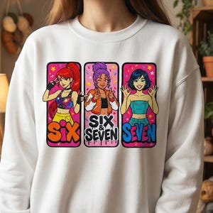 以下が含まれることがあります： 白いスウェットシャツ。3人の漫画の女性のカラフルなグラフィックと「SIX & SEVEN」の文字が特徴です。女性はさまざまなポーズと衣装で、オレンジ、紫、青などの鮮やかな色を使用しています。スウェットシャツはカジュアルなスタイルです。