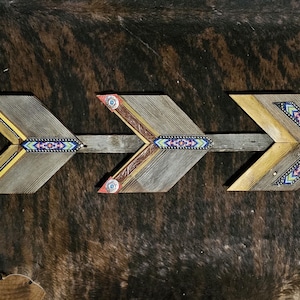 以下が含まれることがあります： 素朴なデザインの木製の矢が3本。矢はグレーと黄色の配色で、青、ピンク、緑の装飾的なビーズアクセントが付いています。矢は茶色の背景に対して水平に配置されています。