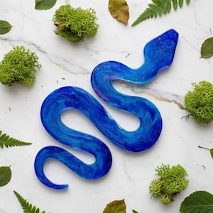May include: A vibrant blue resin snake sculpture with swirling patterns. The snake is surrounded by green moss and leaves on a white marble surface. The sculpture's design is detailed and eye-catching, perfect for home decor.