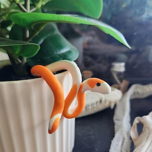 May include: A handcrafted orange and white snake figurine, coiled around the rim of a white ceramic pot. The snake is a decorative item, possibly a plant accessory, with a detailed head and body. Green plant leaves are visible in the background.