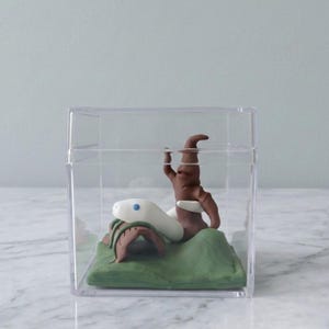 May include: A whimsical diorama inside a clear plastic box. It features a white snake with blue eyes, a brown tree-like structure, and green and brown clay foliage. The scene is set on a green base, creating a miniature, artistic display.