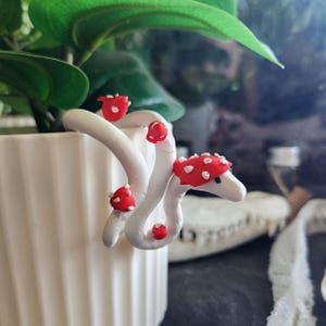 May include: A white, whimsical clay sculpture of a snake-like creature with red mushroom caps. The sculpture is placed on a white pot with green leaves in the background. The mushrooms have white spots.