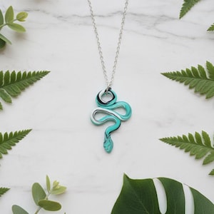 May include: A silver chain necklace with a pendant shaped like a snake. The snake pendant is a swirl of turquoise, white, and black. The necklace is displayed on a white marble surface with green foliage.