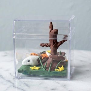 May include: A miniature diorama inside a clear plastic cube. The scene includes a brown tree, a white and brown snake, green grass, and yellow and orange leaves. The diorama is a whimsical, handcrafted art piece.