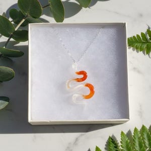 May include: A clear and orange snake pendant necklace on a silver chain, presented in a white box. The snake design has a swirling pattern of clear and orange resin. The necklace is displayed against a soft, neutral background with greenery.