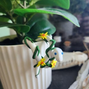 May include: A white ceramic pot with a green plant and a decorative white snake figurine with blue eyes, yellow flowers, and green vines. The figurine is wrapped around the edge of the pot.