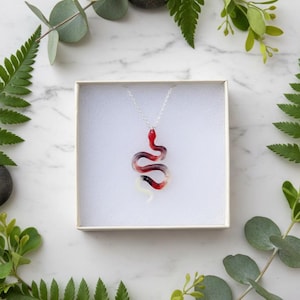 May include: A silver chain necklace with a pendant shaped like a snake. The snake pendant is red, white, and purple, and is displayed in a white box. Green foliage surrounds the box on a marble surface.