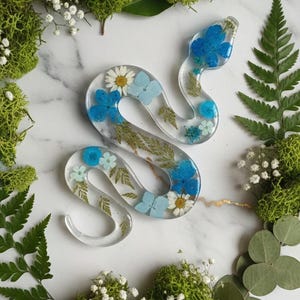 May include: A clear resin snake sculpture filled with blue and white flowers and greenery. The snake is surrounded by green moss, ferns, and small white flowers, all arranged on a white marble surface.