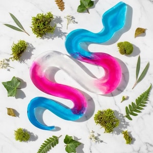 May include: A colorful, abstract snake sculpture in shades of blue, pink, and white. The resin art piece is surrounded by greenery, including moss, leaves, and small white flowers, arranged on a white marble surface.