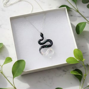 May include: A silver necklace with a snake pendant. The snake is black with green and white accents. The pendant is inside a white box, with green leaves in the background.