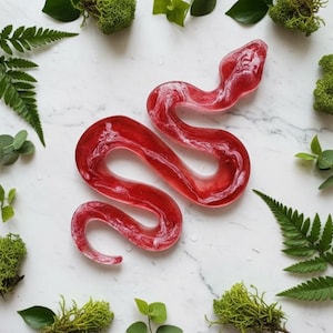 May include: A red and white swirled resin snake figurine. The snake is in a coiled position, with a detailed head and body. The figurine is surrounded by green moss and ferns on a white marble surface.
