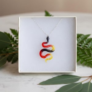 May include: A silver necklace featuring a pendant in the shape of a snake. The snake pendant is colored in red, black, and yellow. The necklace is displayed inside a white box.