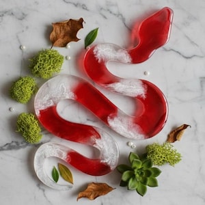 May include: A red and white resin snake sculpture with a marbled effect, surrounded by green moss, leaves, and pearls. The snake is a decorative art piece, possibly a paperweight or home decor item.