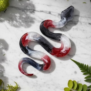 May include: A decorative snake figurine in a winding pose. The sculpture features a blend of colors, including red, black, white, and gray, creating a marbled effect. The snake is placed on a white marble surface.
