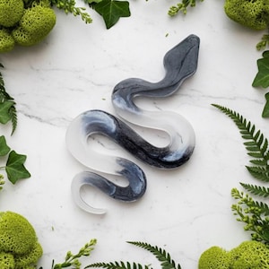 May include: A decorative snake figurine with a gradient design, transitioning from a dark gray head to a white tail. The snake is surrounded by green foliage on a white marble surface.
