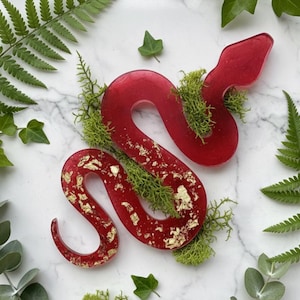 May include: A red resin snake sculpture with gold leaf accents and green moss. The snake is coiled and surrounded by ferns and greenery, creating a naturalistic aesthetic. The sculpture is on a white marble surface.