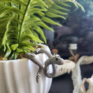 May include: A gray, coiled snake figurine with white spots and small white insects, wrapped around the edge of a white ceramic pot. The pot contains a green fern plant. The background includes a skull and other decorative elements.