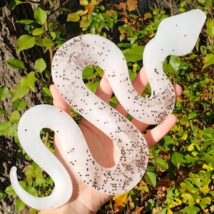 May include: A white, glitter-filled snake-shaped decorative item. The snake is made of a translucent material and is filled with small, dark glitter particles. The snake is held against a backdrop of green foliage.