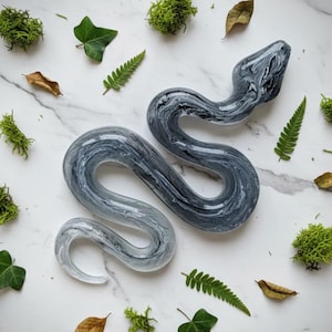 May include: A decorative snake sculpture with a marbled gray and white design. The snake is surrounded by green moss, leaves, and ferns on a white marble surface. The sculpture has a smooth, glossy finish.