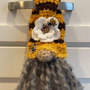 May include: A crocheted gnome towel topper with a yellow and brown striped hat, a white crocheted flower with a button center, and two small bee embellishments. The gnome has a gray yarn beard.