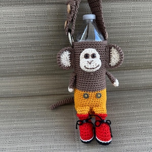 May include: A crocheted monkey water bottle holder. The monkey has a brown body, tan ears, and a white face. It wears yellow shorts with brown buttons and red shoes. A brown strap is attached to the top for carrying.