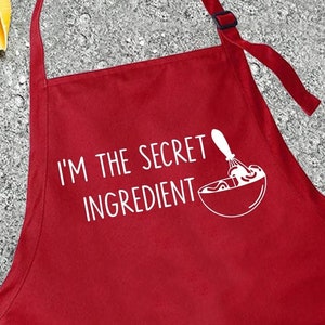 May include: A red apron with the text "I'M THE SECRET INGREDIENT" in white. The apron has a pocket and adjustable neck strap. Various ingredients like tomatoes, pasta, and olives are arranged around the apron.
